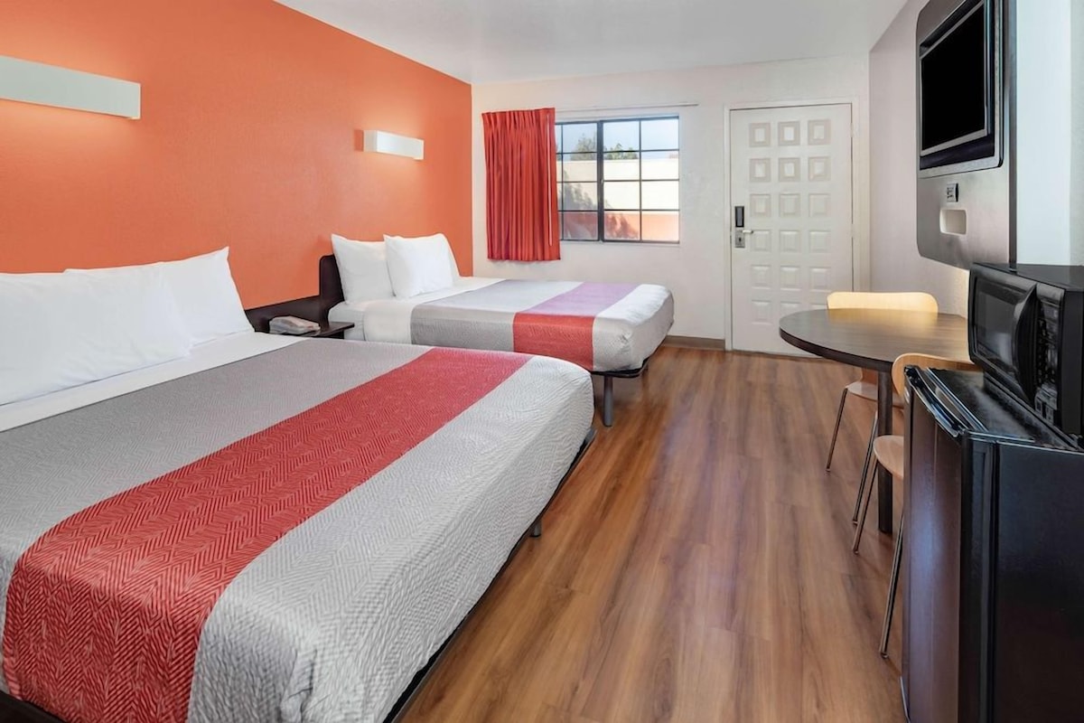 A hotel room with two beds, a TV, a small table, and a window with curtains.