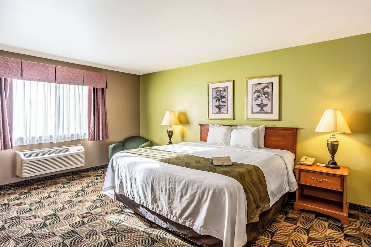 A hotel room with a bed, two bedside lamps, a nightstand, a chair, a window with curtains, and a wall-mounted air conditioner.