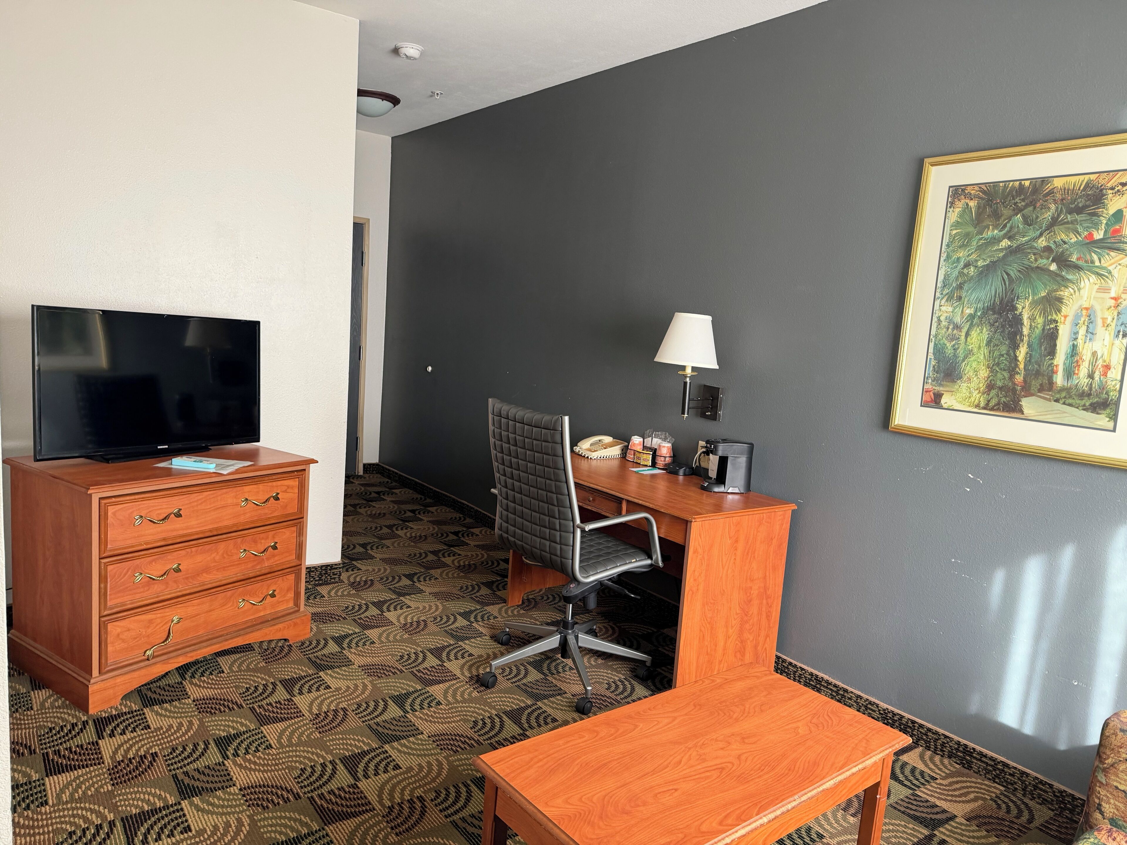 Une chambre d’hôtel avec un téléviseur posé sur une commode en bois, un bureau avec une chaise, une lampe et un tableau encadré accroché au mur.