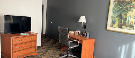 Une chambre d’hôtel avec un téléviseur posé sur une commode en bois, un bureau avec une chaise, une lampe et un tableau encadré accroché au mur.