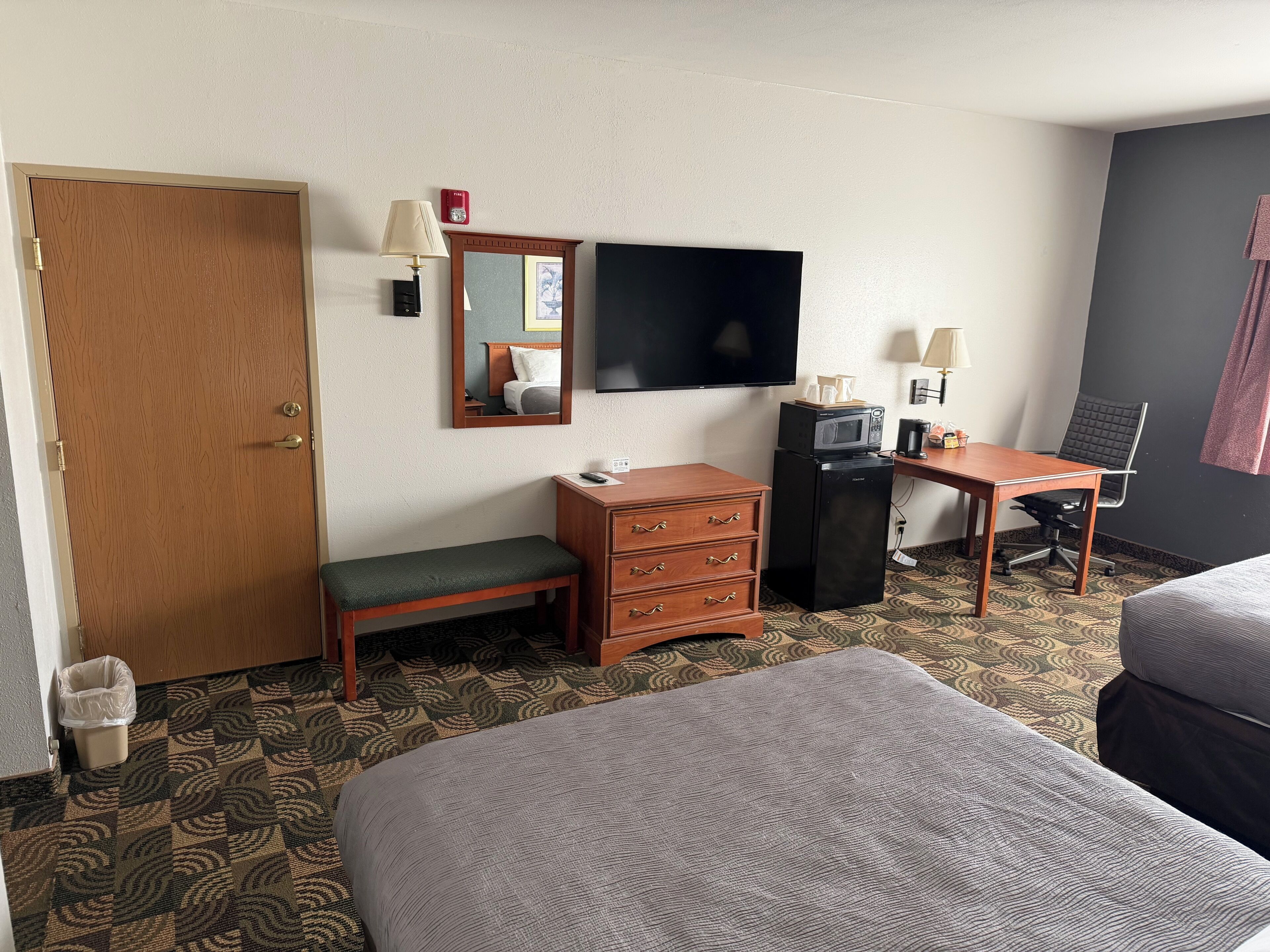 Quarto de hotel com porta de madeira, um banco, escrivaninha com computador, televisão e duas camas.