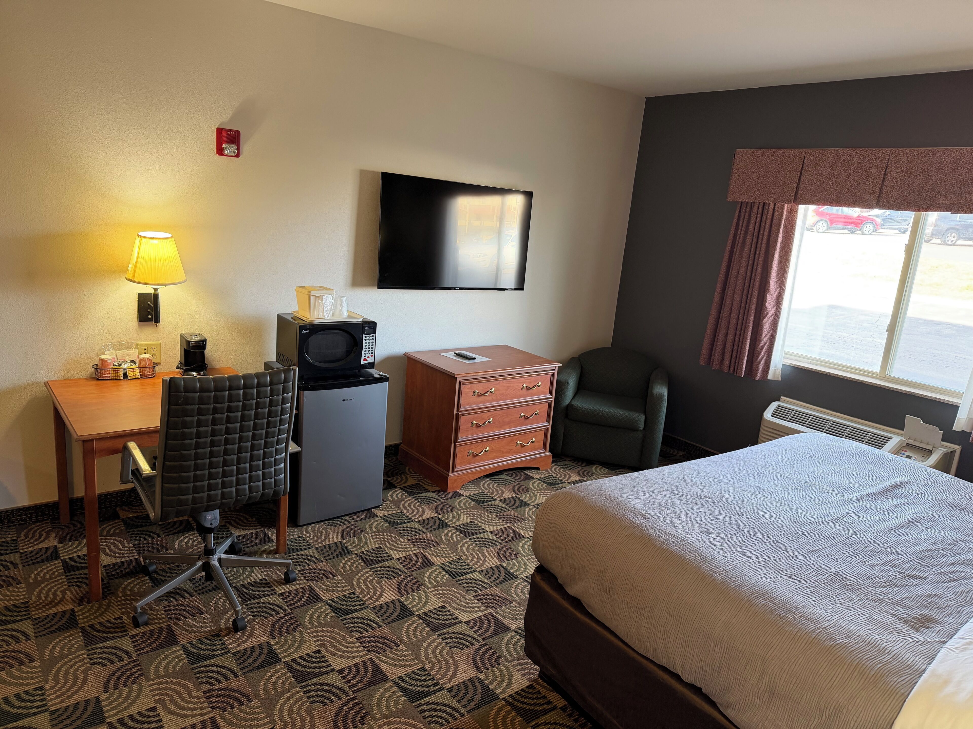 Habitación de hotel con cama, escritorio, silla, microondas, refrigerador, televisor y ventana con cortinas.