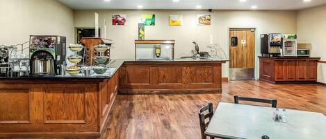 A hotel lobby with a wooden reception desk, a coffee machine, a microwave, and a seating area with tables and chairs.
