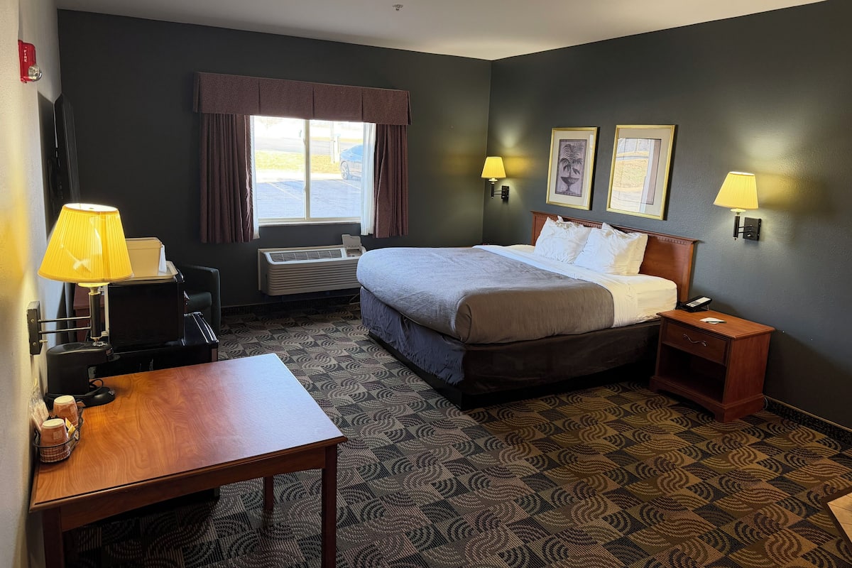 A hotel room with a bed, bedside tables, a desk, a chair, a window with curtains, wall-mounted lamps, and a wall-mounted air conditioner.