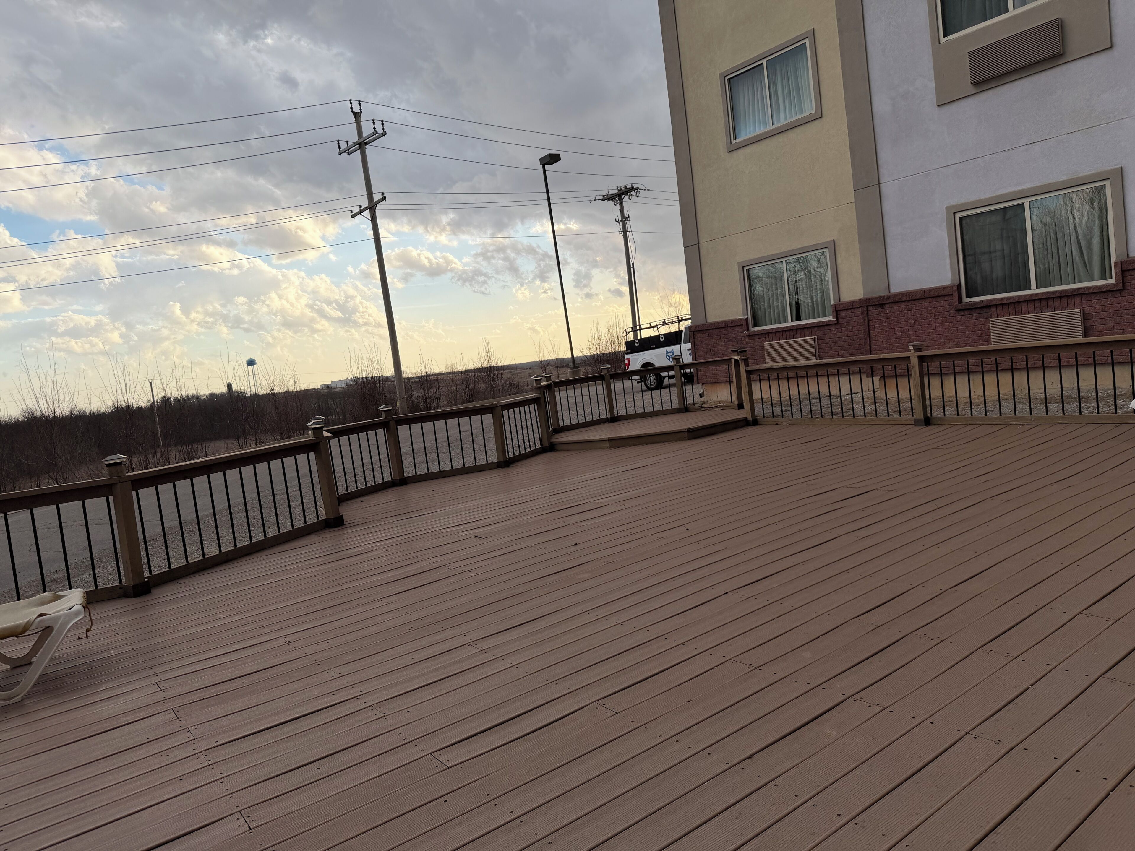 A wooden deck with railings, a white truck parked nearby, and a building with windows and a brick wall in the background.