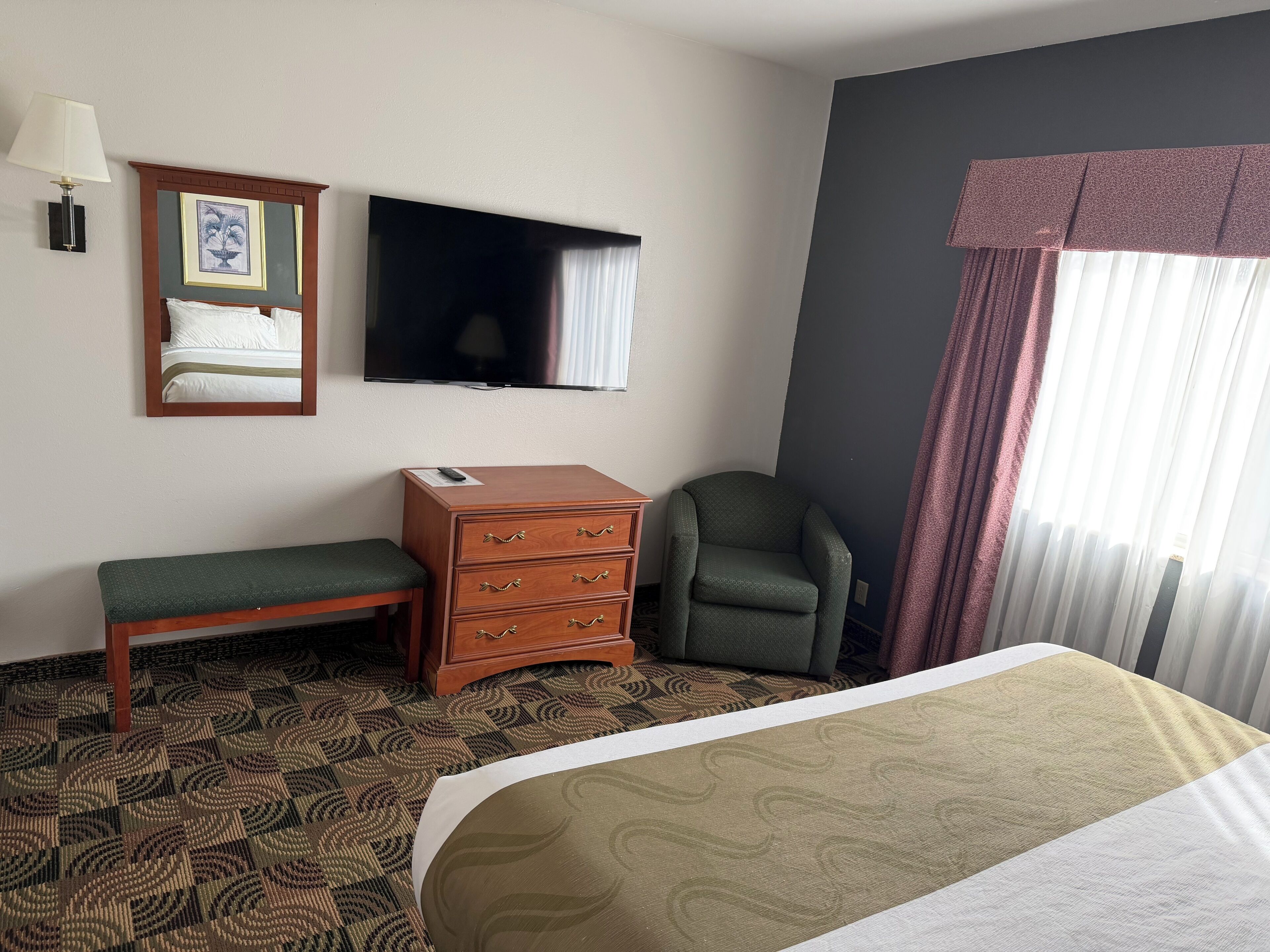 Une chambre d’hôtel avec un lit, une commode en bois, une chaise, un téléviseur et une fenêtre avec des rideaux.