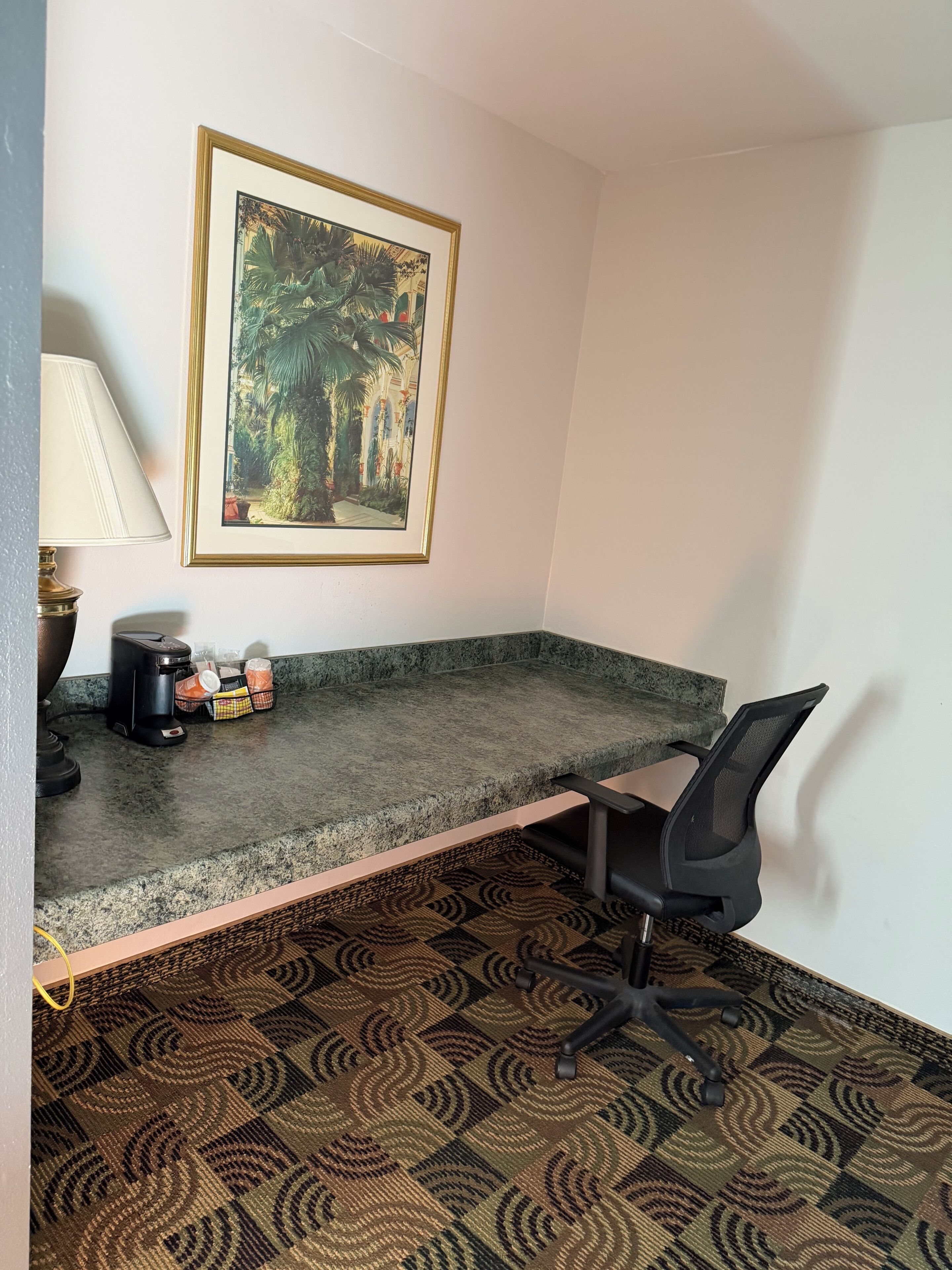 A corner office with a desk, chair, coffee maker, and a framed picture on the wall.
