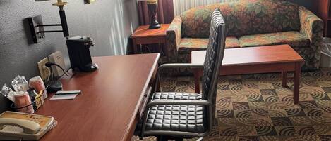 A hotel room with a desk, chair, sofa, and a patterned carpet.
