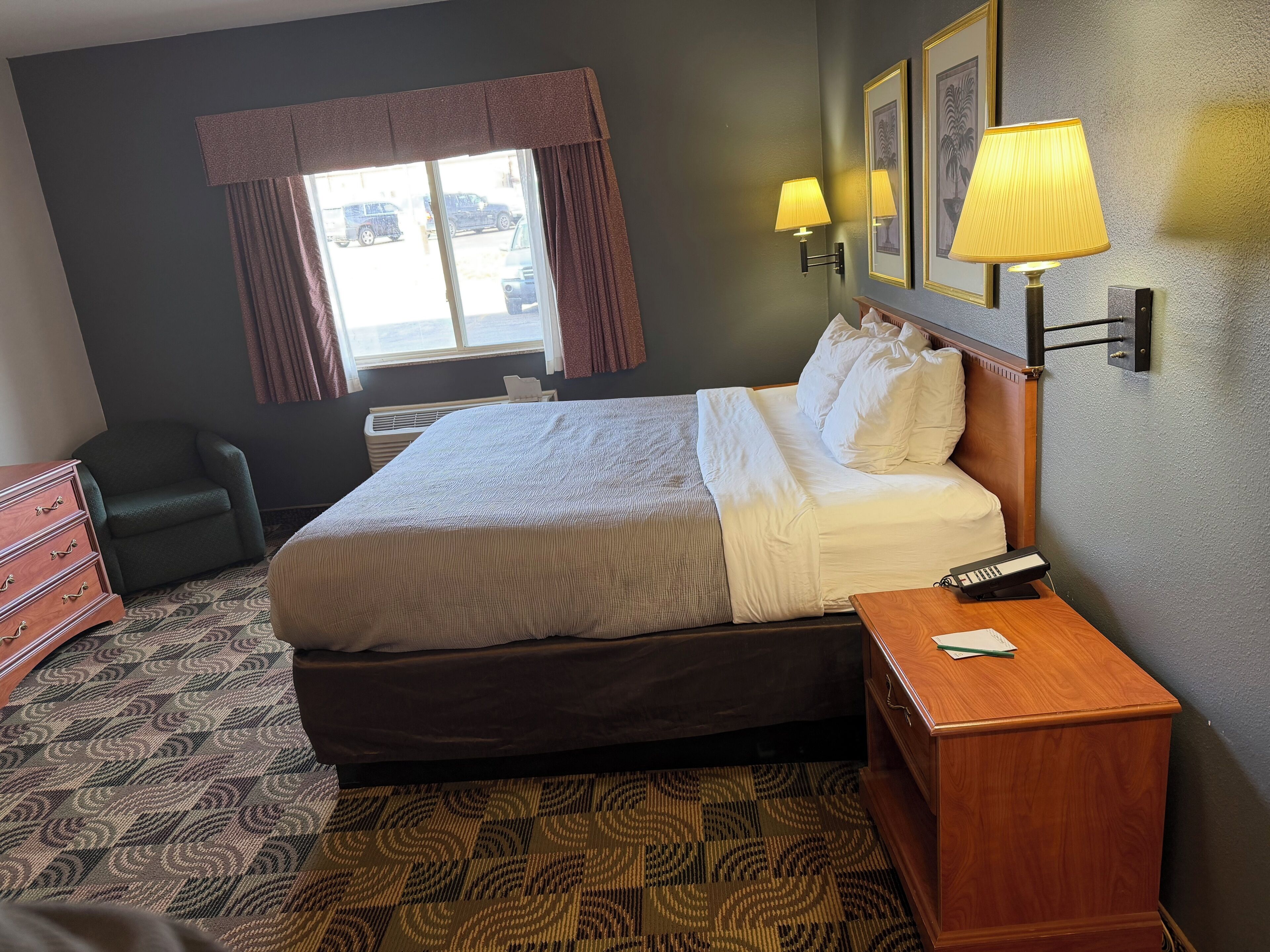 A hotel room with a bed, a desk with a lamp, a chair, a dresser, and a window with curtains.
