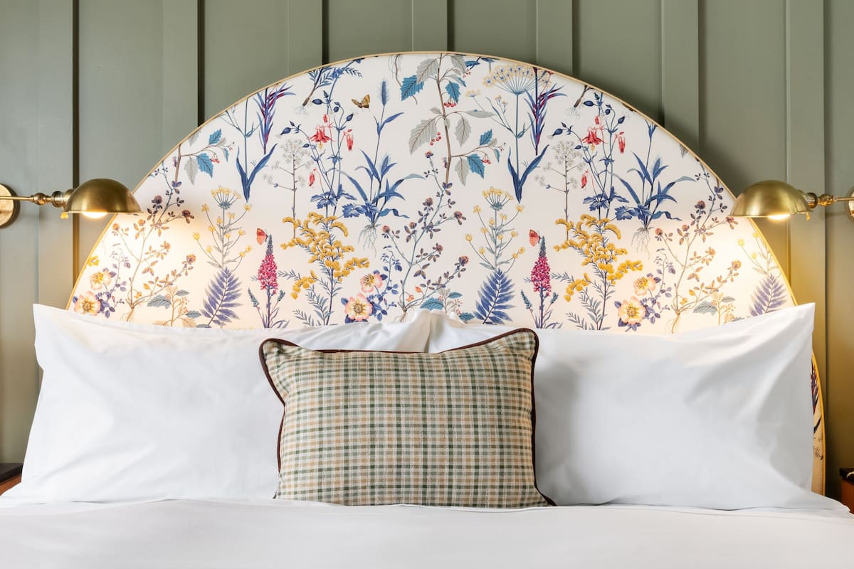 Una cama con cabecera floral, ropa de cama blanca y almohada de cuadros.