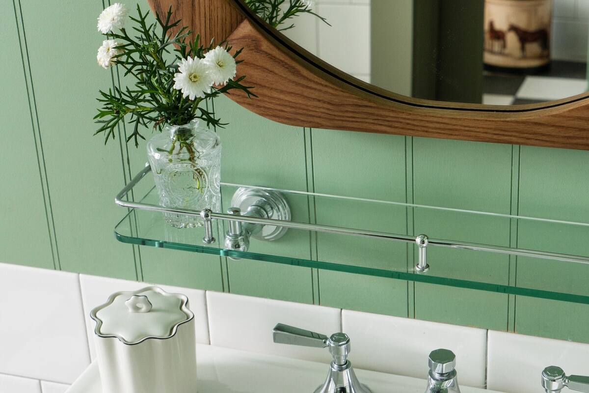 Un baño con un lavamanos blanco, grifos cromados y un estante de vidrio con un florero de flores blancas.