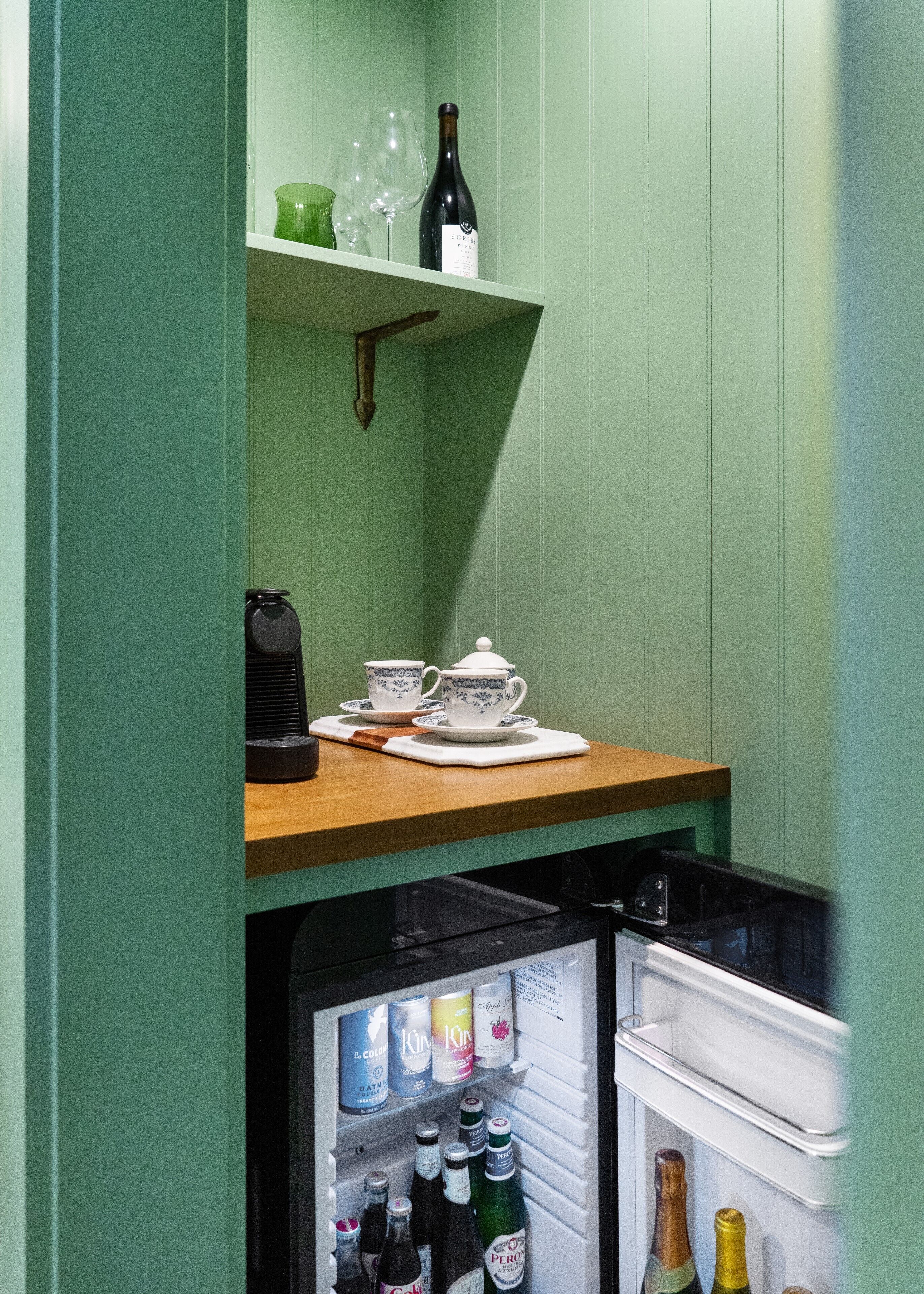 A compact kitchen with a wooden countertop, a small fridge, and a shelf with a wine bottle and glasses.