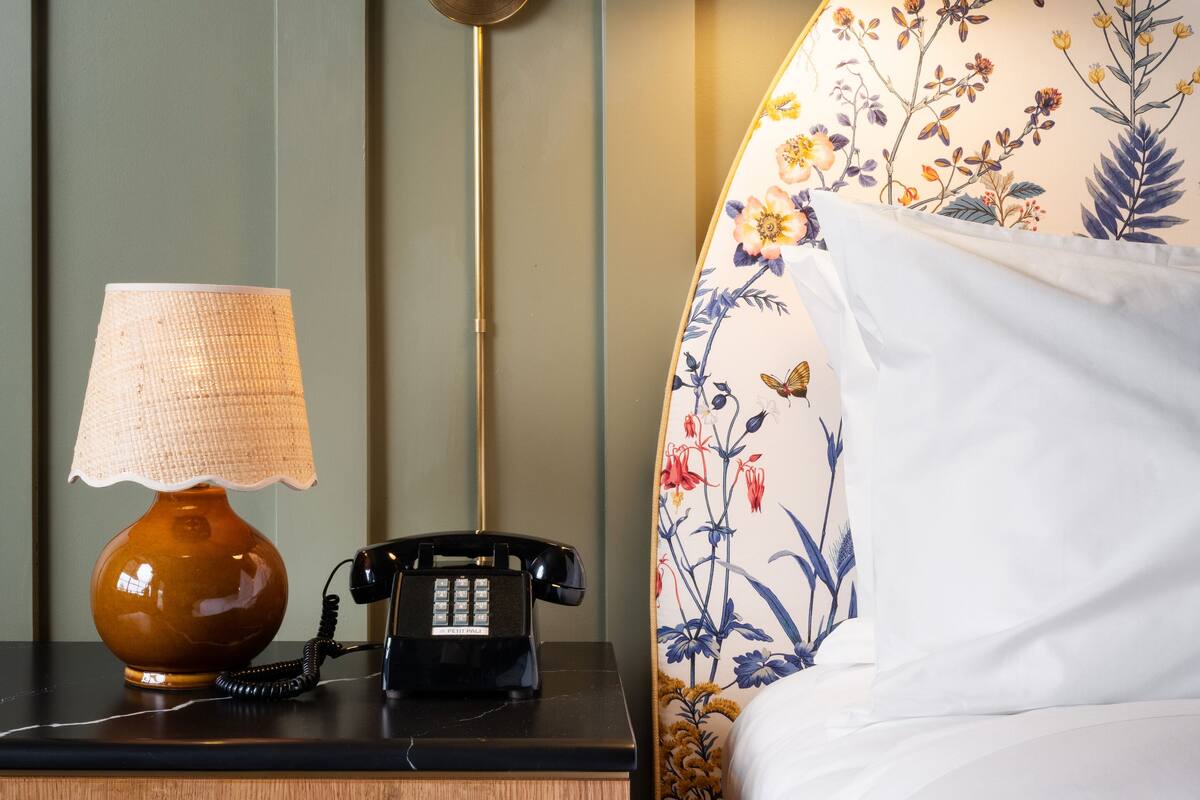 Una mesita de noche de madera con una lámpara, un teléfono y una cama con ropa de cama de estampado floral.