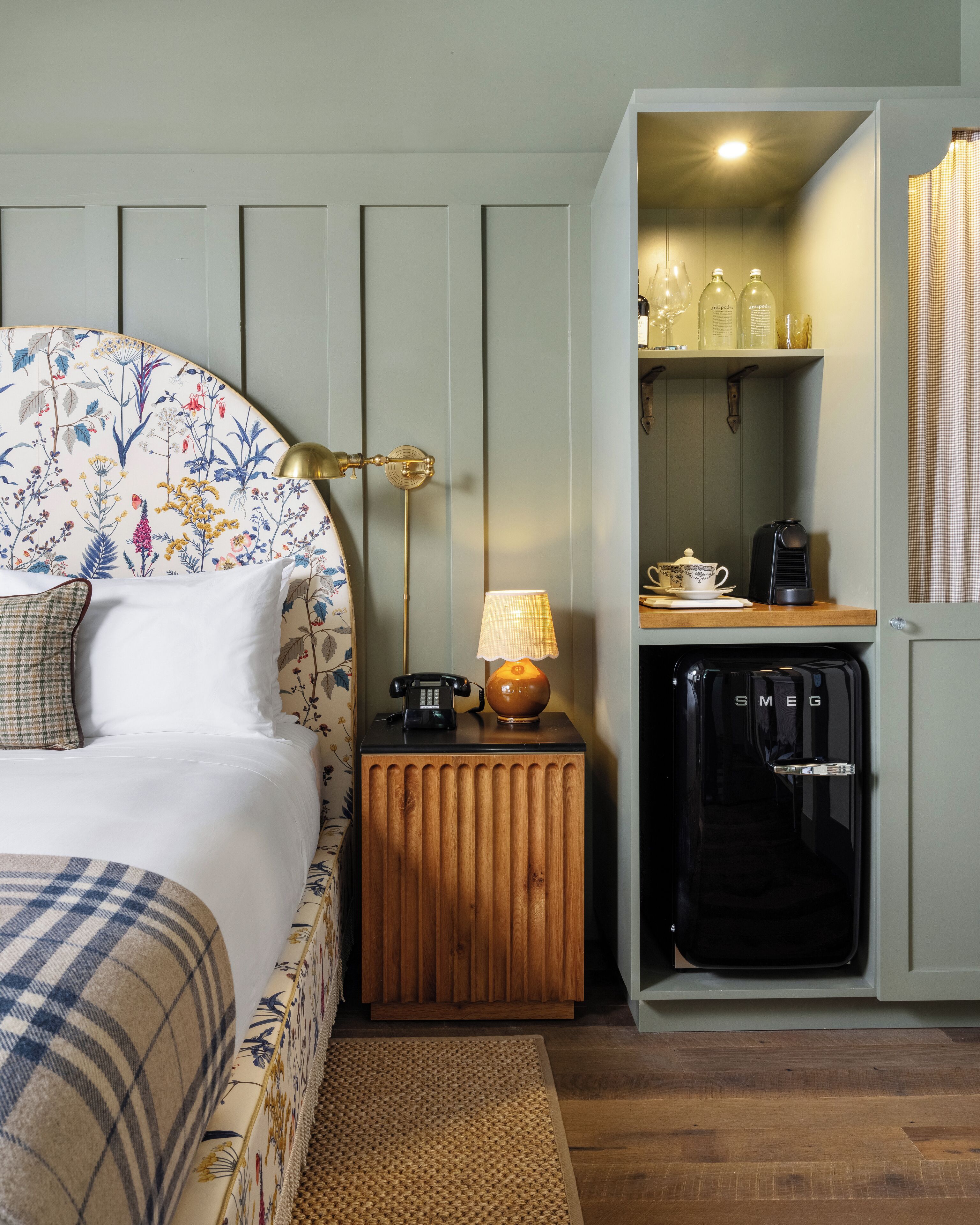 A bedroom with a floral headboard, a bedside lamp, a telephone, and a minibar with a coffee maker.