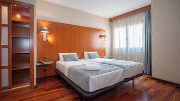 Habitación de hotel con armario de madera, dos camas con ropa blanca, mesita de noche, lámpara y ventana con cortinas.
