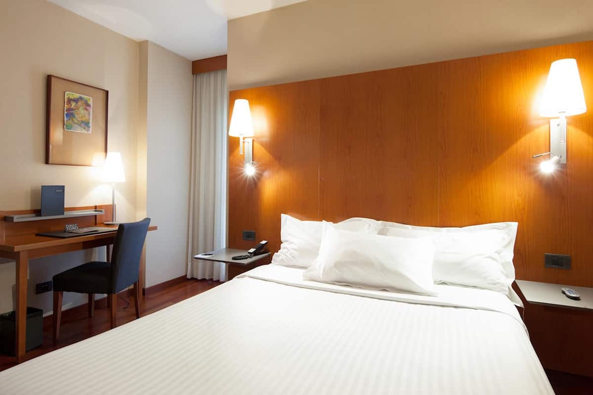 Hotellihuone, jossa on sänky, työpöytä, tuoli ja kaksi seinään kiinnitettyä lamppua.