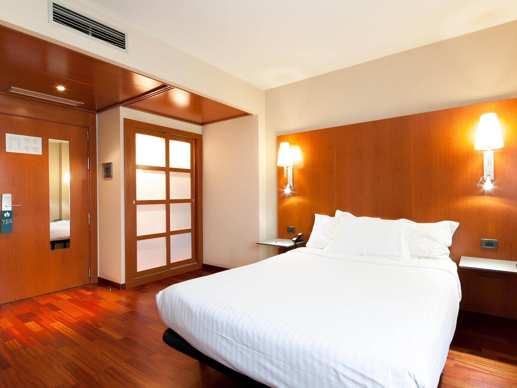 Hotelkamer met een groot bed, houten lambrisering, een nachtkastje, twee wandlampen en een deur met een geëtste glazen ruit.
