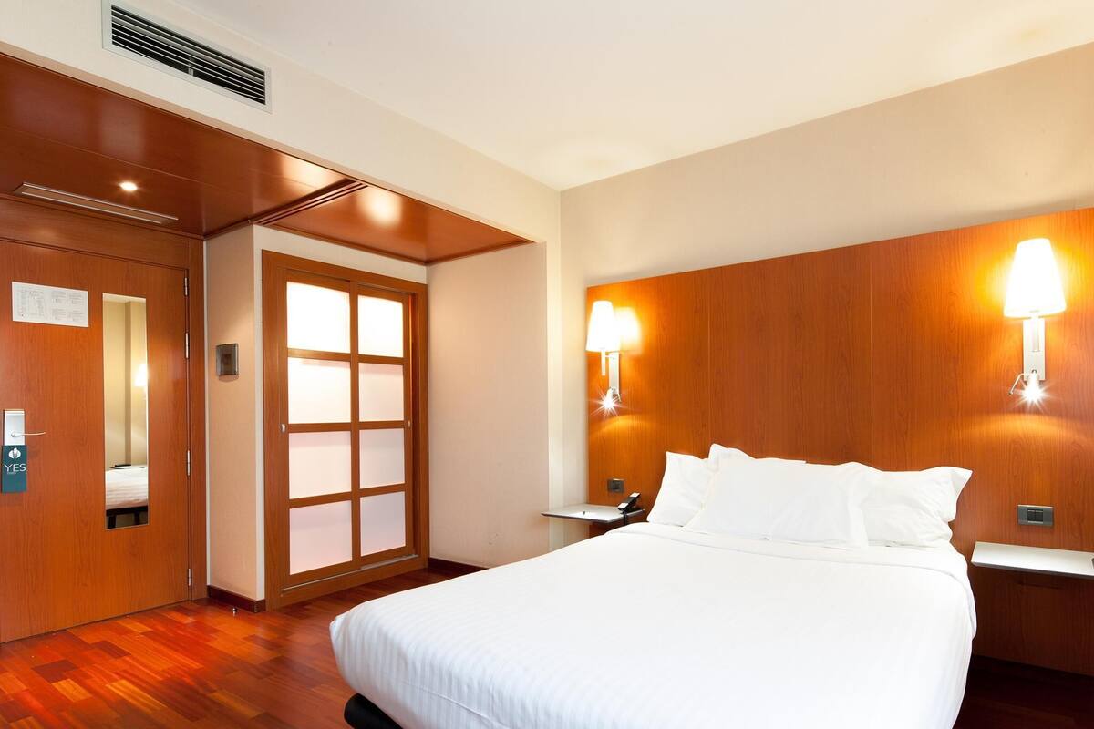 Ein Hotelzimmer mit einem großen Bett, Holzvertäfelung, einem Nachttisch, zwei Wandlampen und einer Tür mit Milchglasfenster.