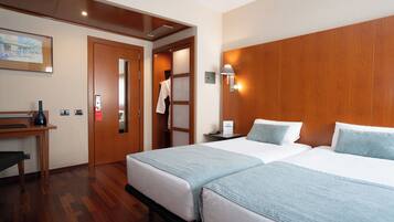 A hotel room with two beds, a desk, a chair, a door, and a wall-mounted lamp.