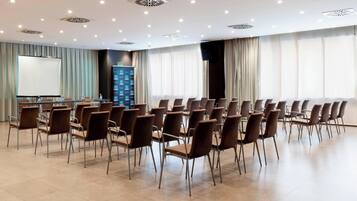 Een vergaderruimte met rijen stoelen, een scherm voor presentaties en een groot raam met gordijnen.