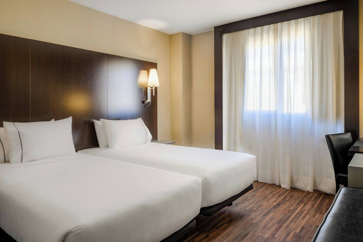 Ein Hotelzimmer mit zwei Betten, einem Schreibtisch, einem Stuhl und einem großen Fenster mit Vorhängen.