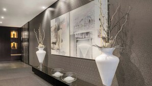 A modern lobby with a dark textured wall, a large abstract painting, and two white vases with branches on a reflective surface. - AC Hotel Elda by Marriott (Elda)