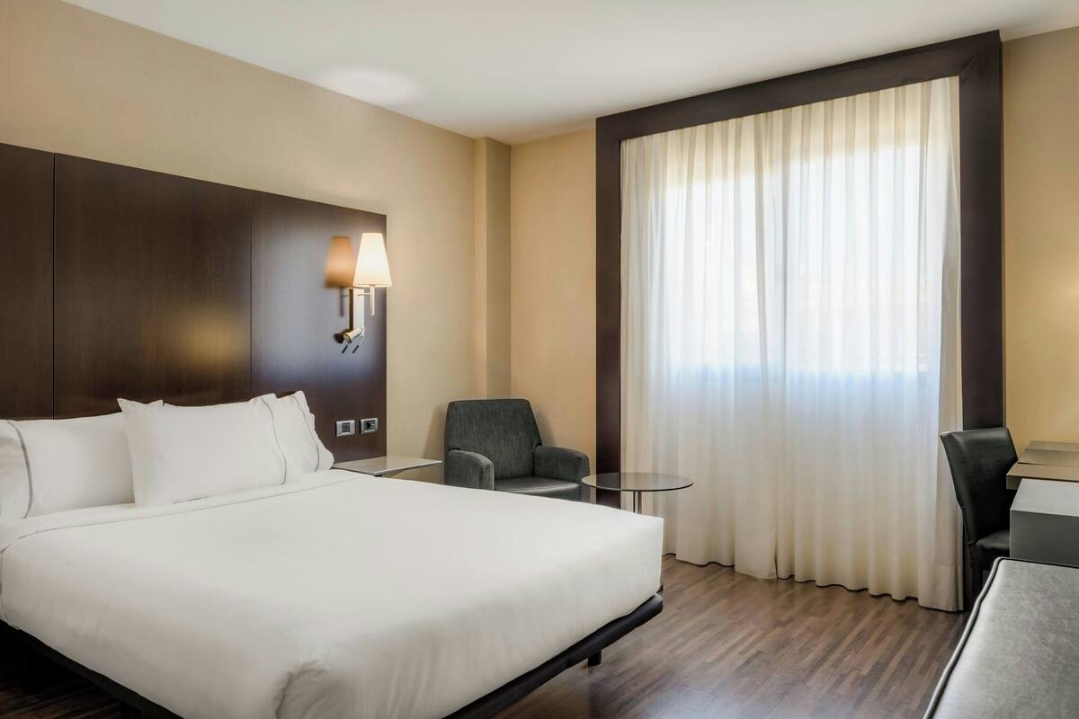 Ein Hotelzimmer mit einem großen Bett, einem Schreibtisch, einem Stuhl, einem kleinen Tisch, einer Lampe und einem Fenster mit Vorhängen.