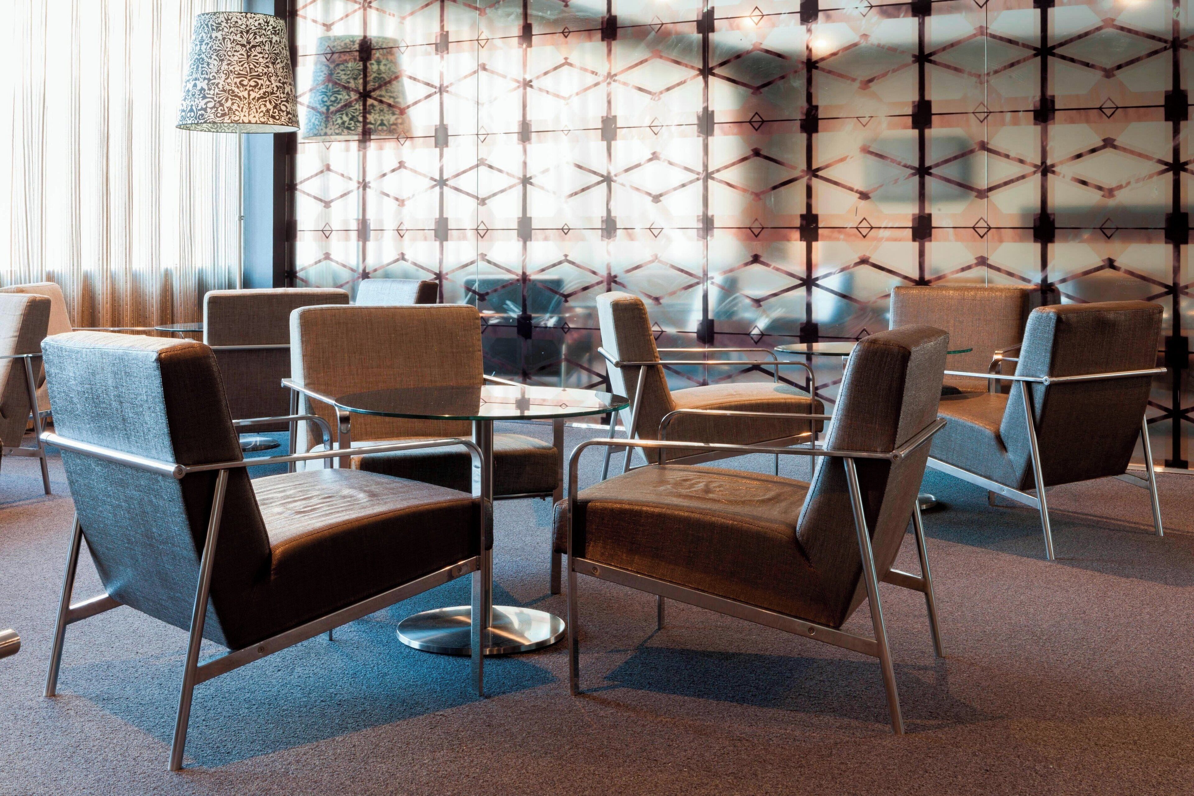 Un área de descanso moderna con sillas contemporáneas y una mesa de vidrio, con un papel tapiz de diseño geométrico y una lámpara colgante.