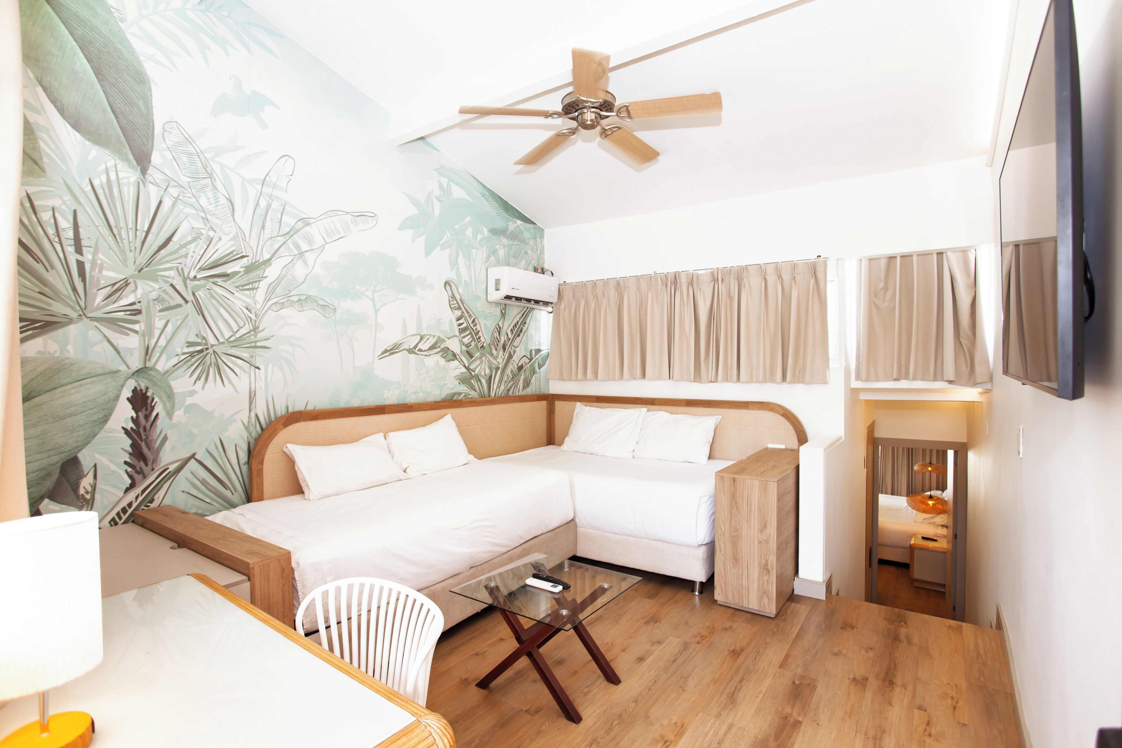 Une chambre d’hôtel moderne dotée d’un canapé d’angle, d’une table basse en verre, d’un ventilateur de plafond, d’un téléviseur à écran plat et d’une fresque représentant des plantes tropicales.