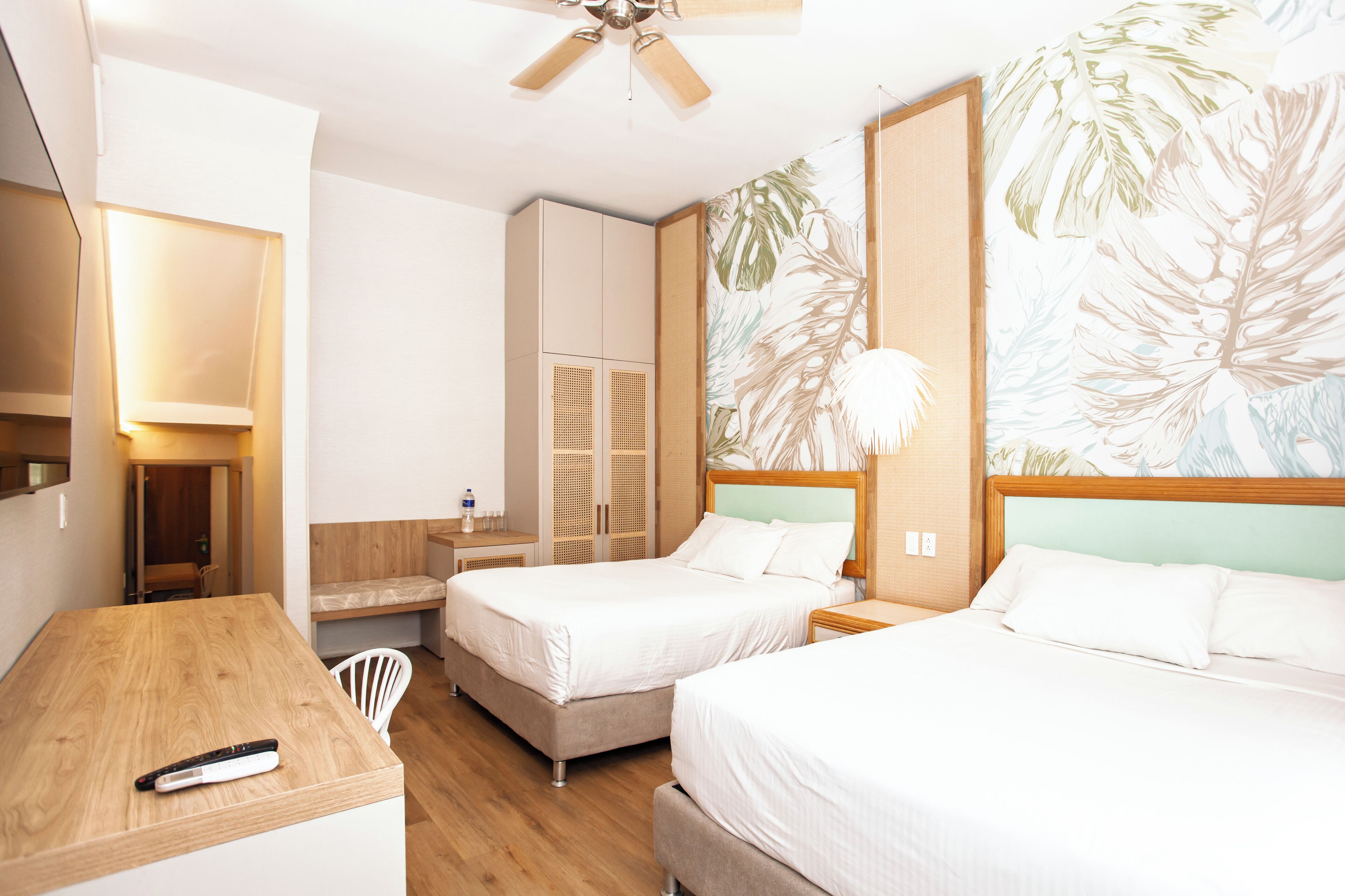 A hotel room with two beds, a wooden desk, a ceiling fan, and a large tropical leaf wallpaper.