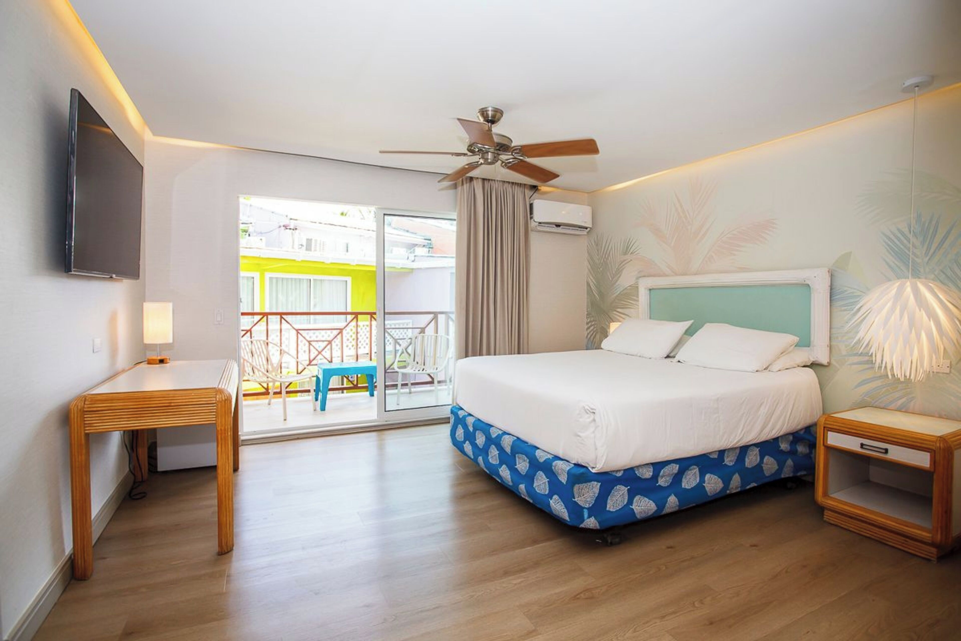 Quarto de hotel com cama, escrivaninha, ventilador de teto, televisão e varanda com vista para um edifício amarelo.