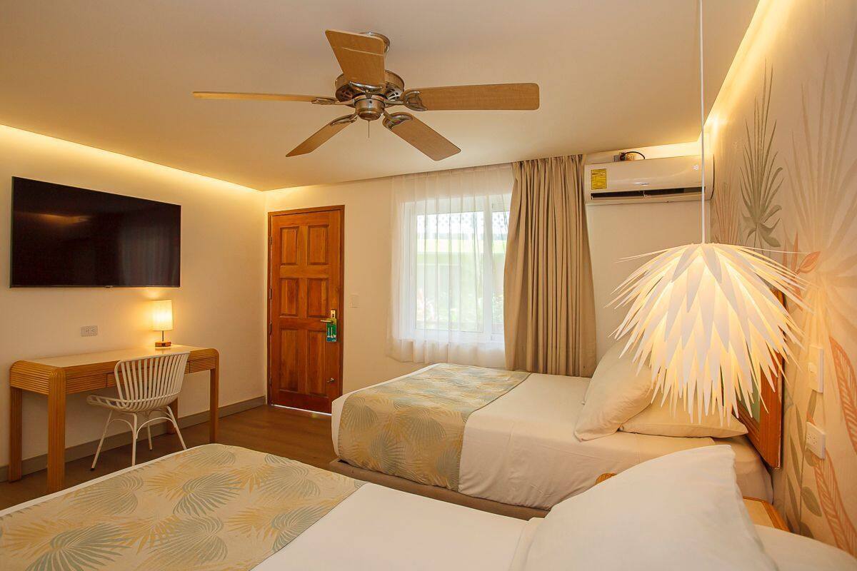 Habitación de hotel con dos camas, un escritorio, una silla, un ventilador de techo, un televisor y una ventana con cortinas.
