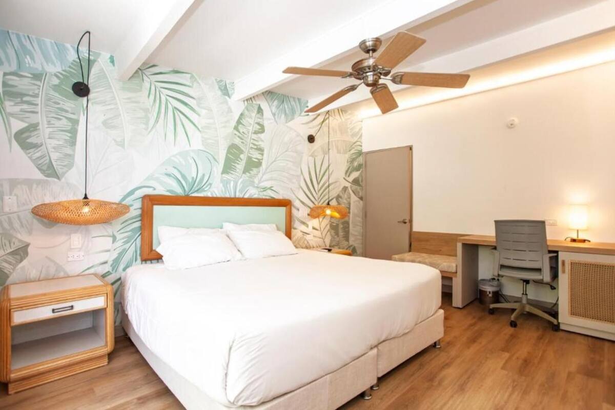 Un dormitorio con cama, escritorio, silla, ventilador de techo y una pared con diseño de hojas.
