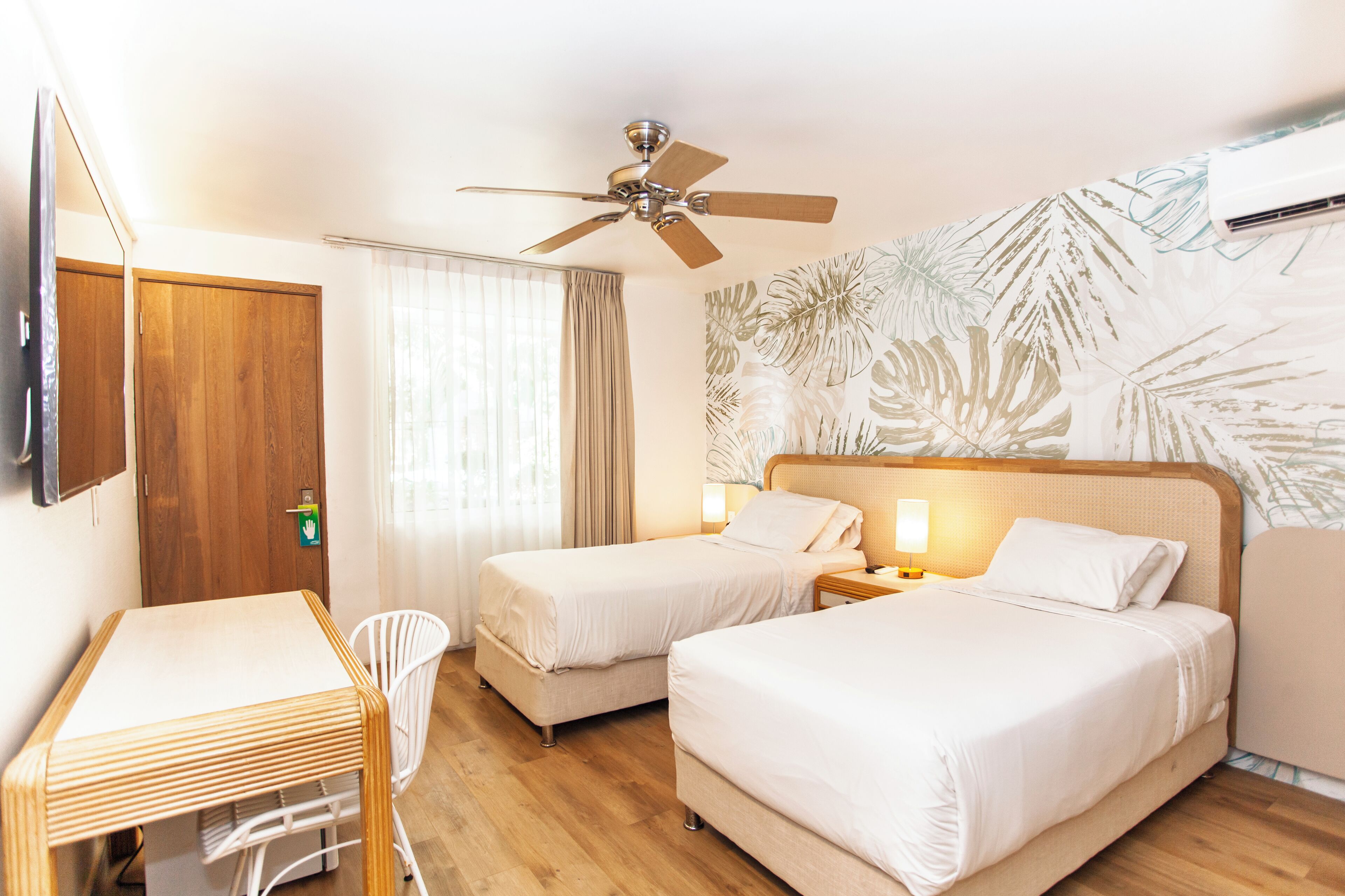 Une chambre d’hôtel avec deux lits, un bureau, une chaise, un ventilateur de plafond et un mur orné d’un motif de feuilles.