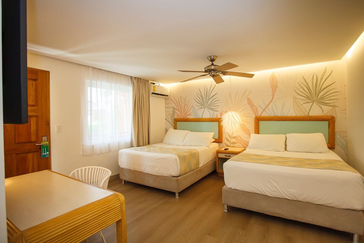 Quarto de hotel com duas camas, um ventilador de teto, uma mesa de madeira e uma janela com cortinas.