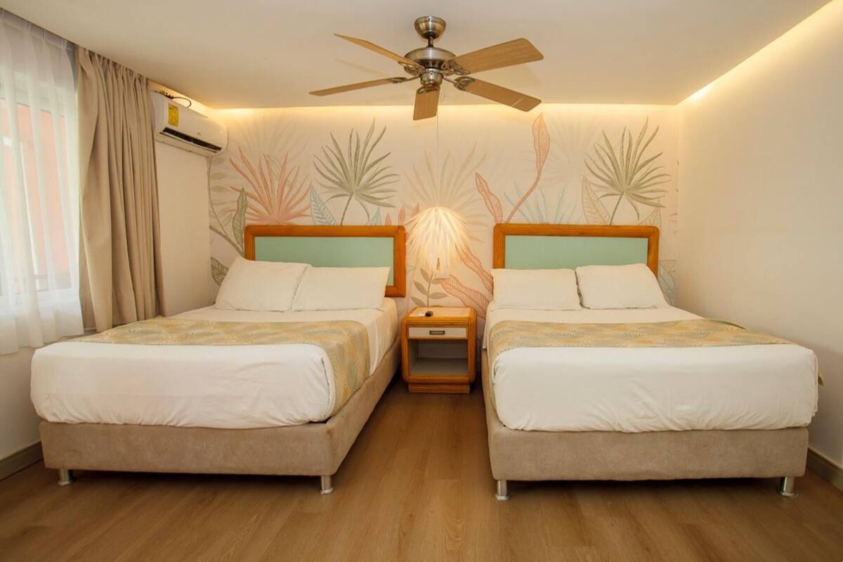 Habitación de hotel con dos camas, un ventilador de techo, una mesita de noche y una pared con diseño de hojas.