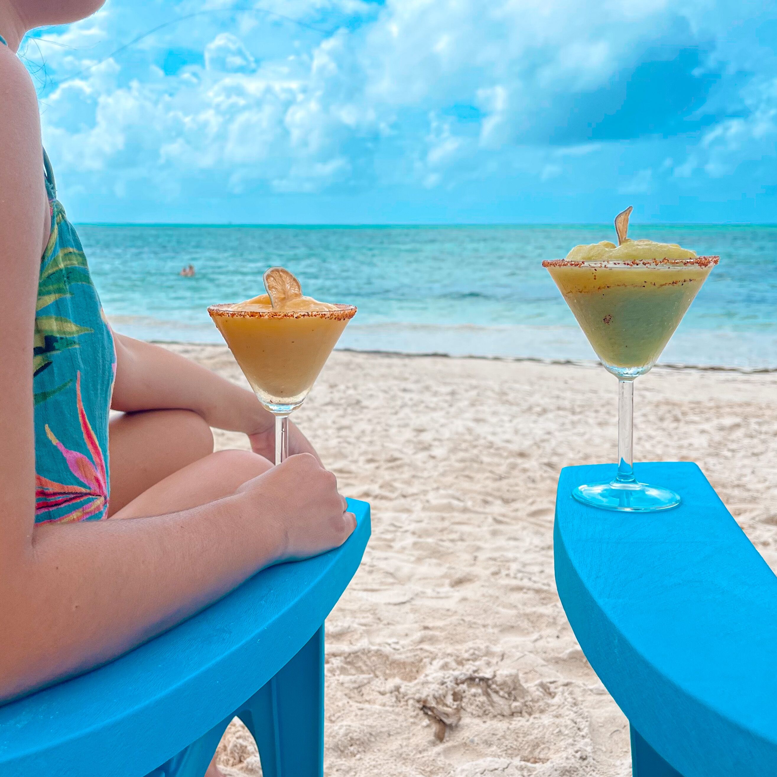 Une personne est assise sur une chaise bleue, sur une plage, et il y a deux boissons sur une table.