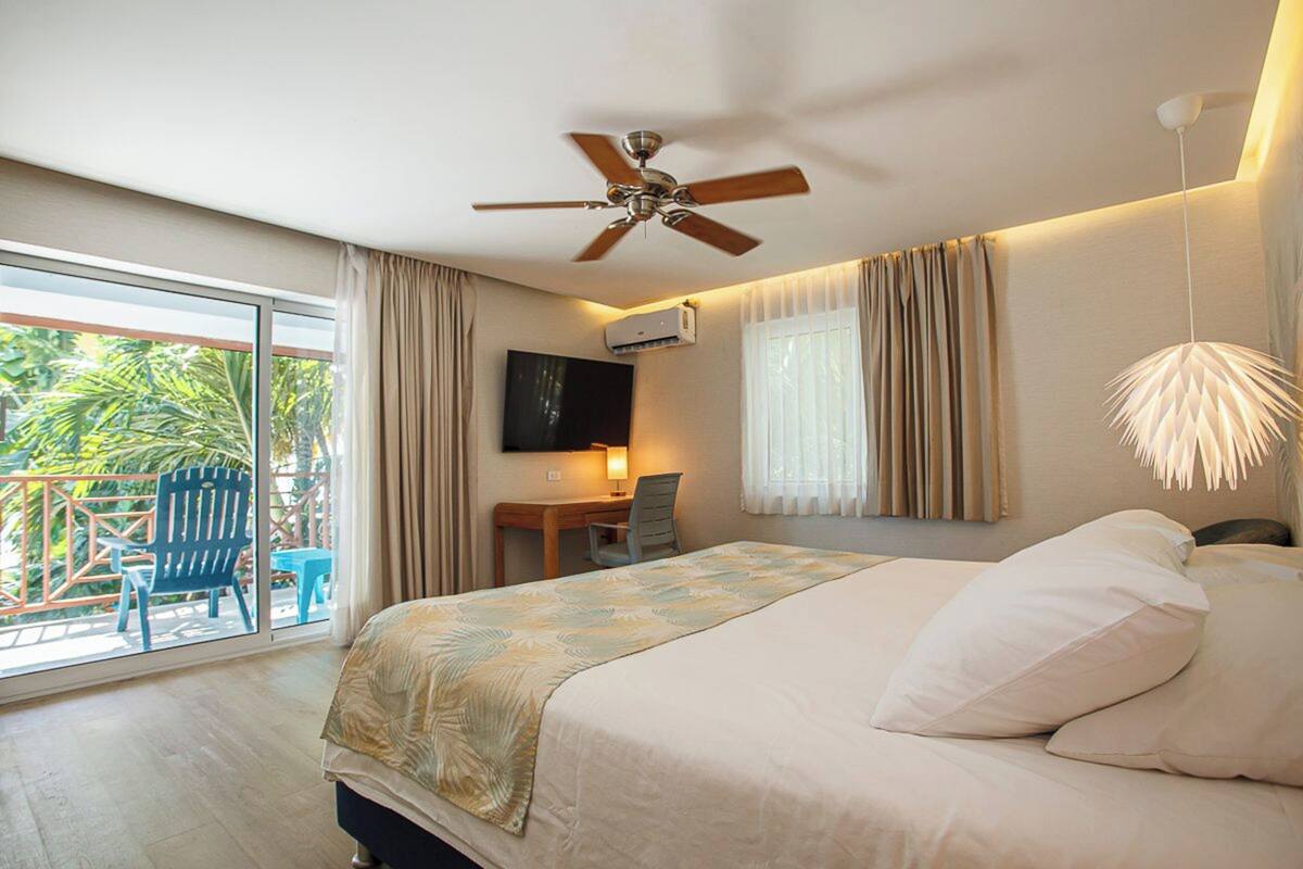 Habitación de hotel con cama, ventilador de techo, televisión, escritorio con silla y balcón con silla y mesa.