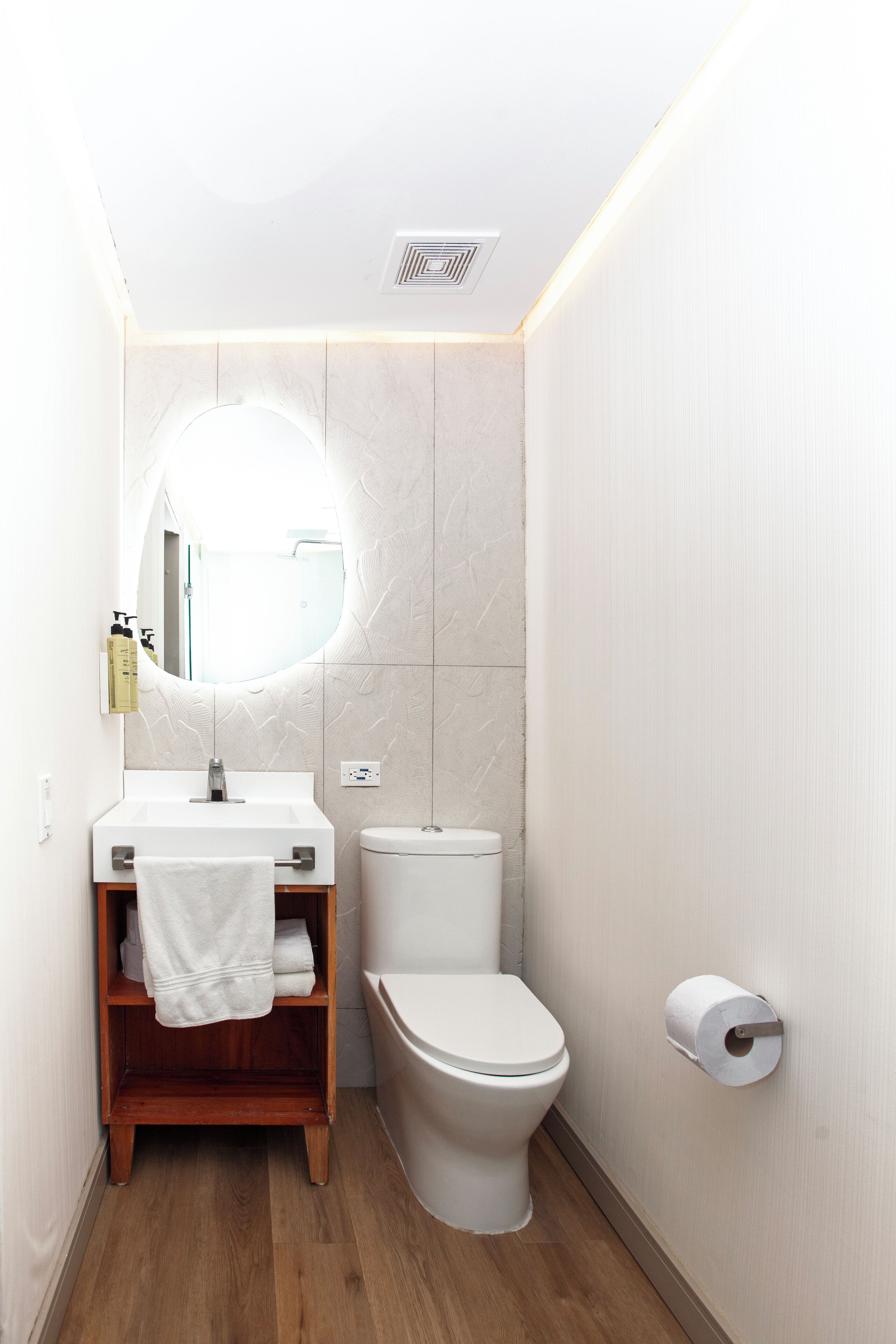 A modern bathroom with a wooden vanity, a round mirror, a toilet, and a toilet paper holder.