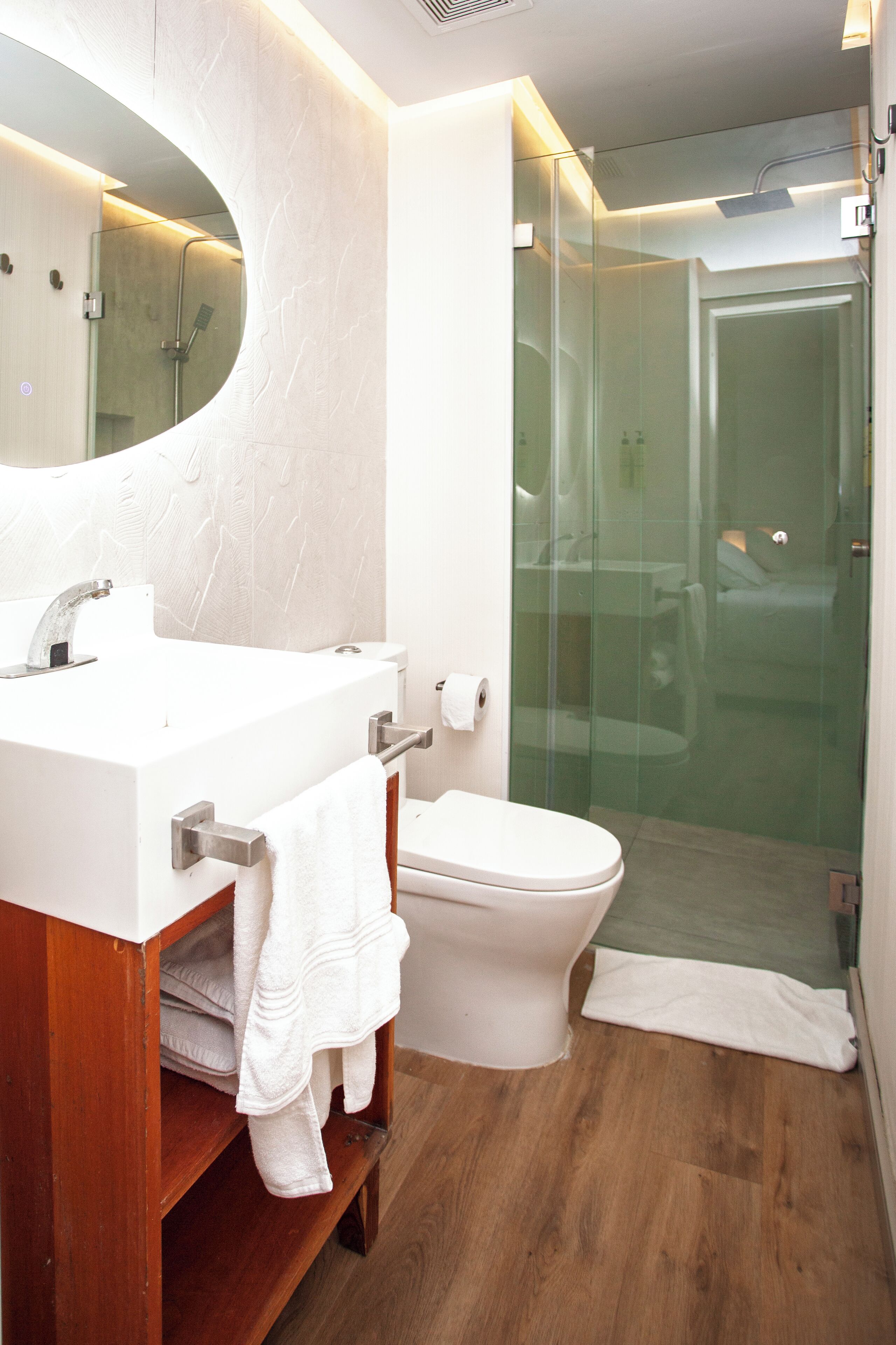 Un baño moderno con tocador de madera, lavamanos blanco, inodoro y ducha con mampara de vidrio.