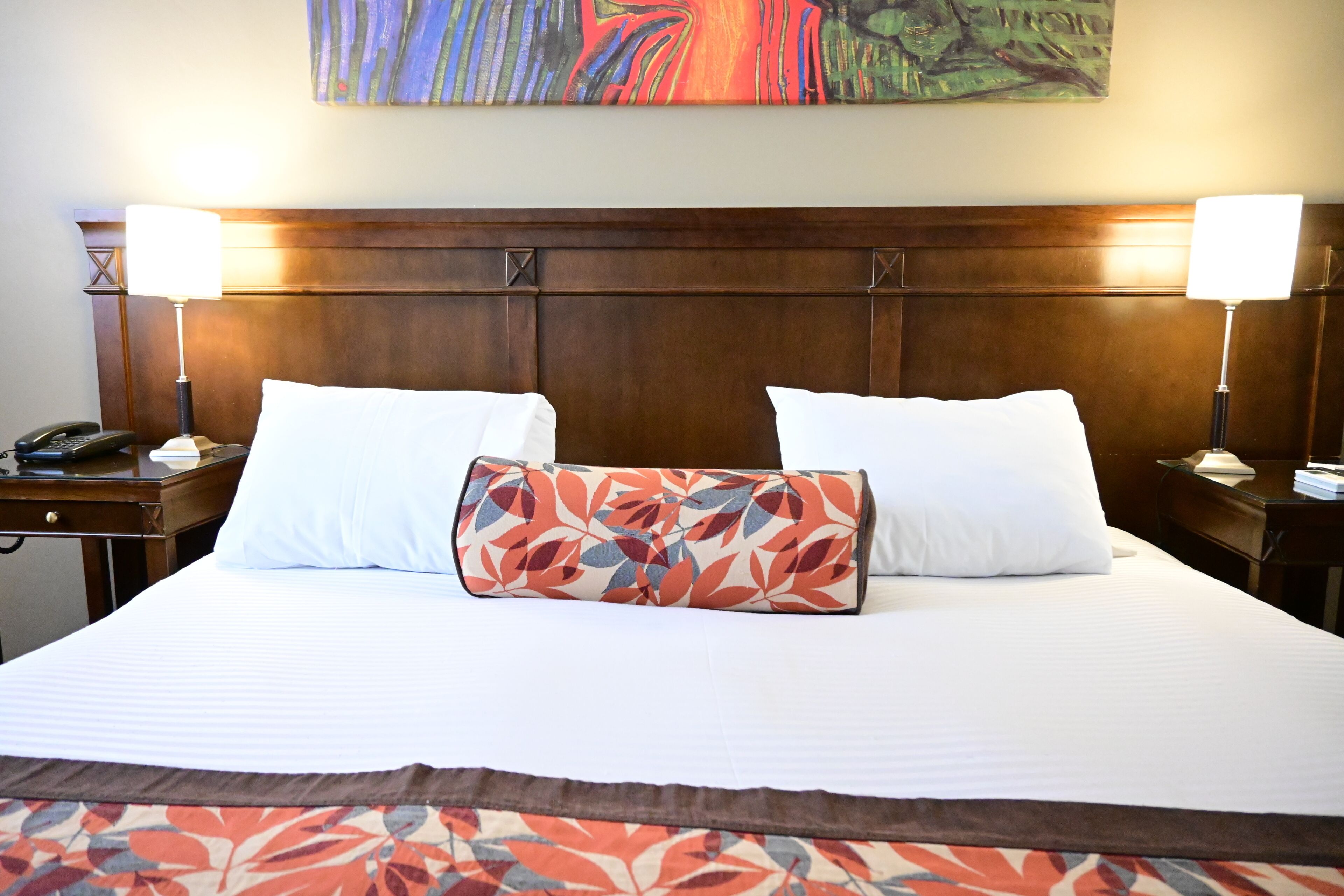 A hotel room with a large bed, two bedside lamps, a telephone, and a colorful painting above the bed.
