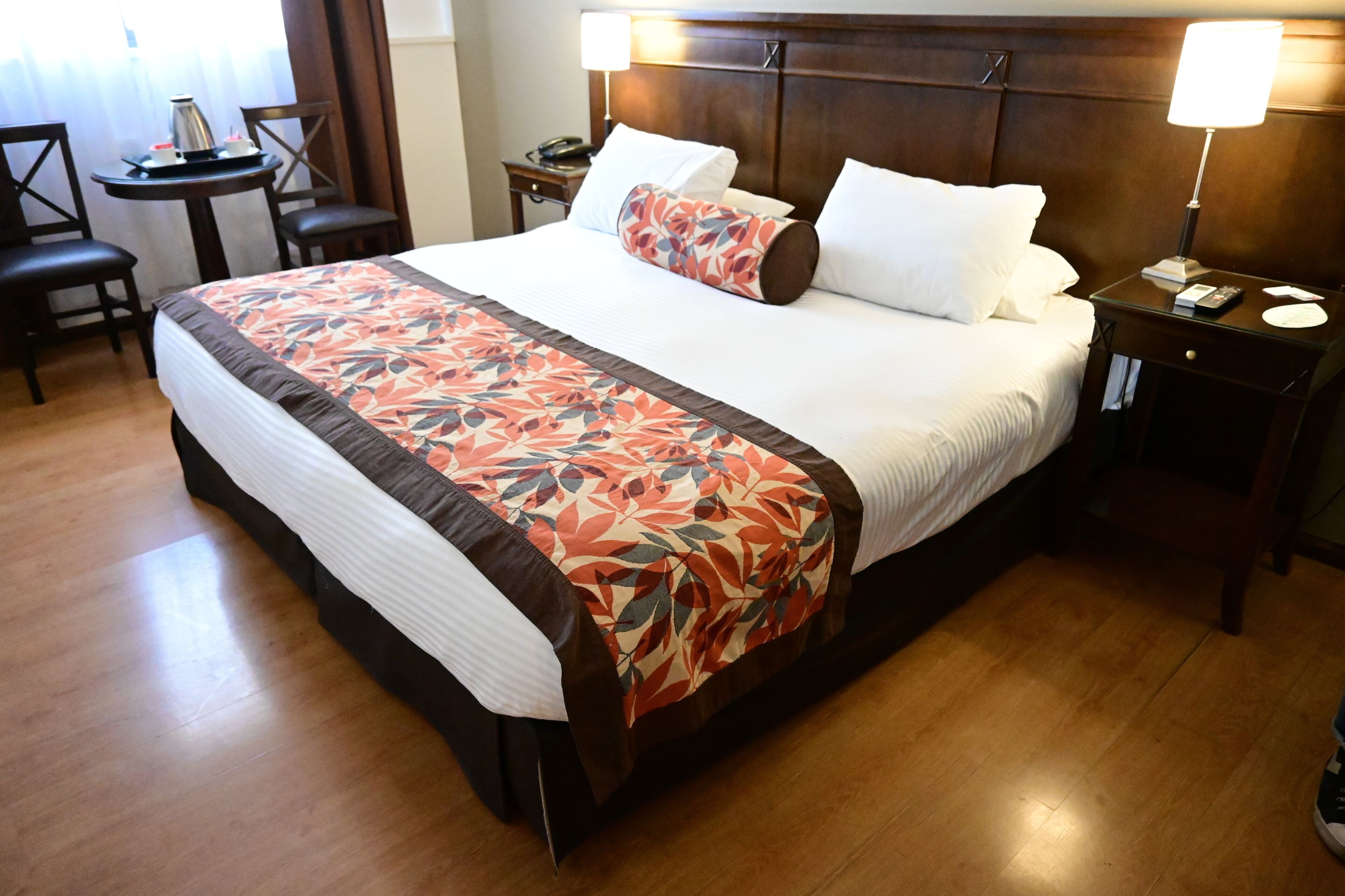 A hotel room with a large bed, a bedside table with a lamp, a small round table with a coffee pot, and two chairs.