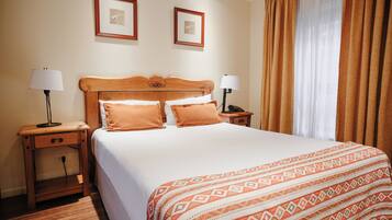 A hotel room with a bed, two bedside tables, a nightstand, a lamp, a telephone, and two framed pictures on the wall.