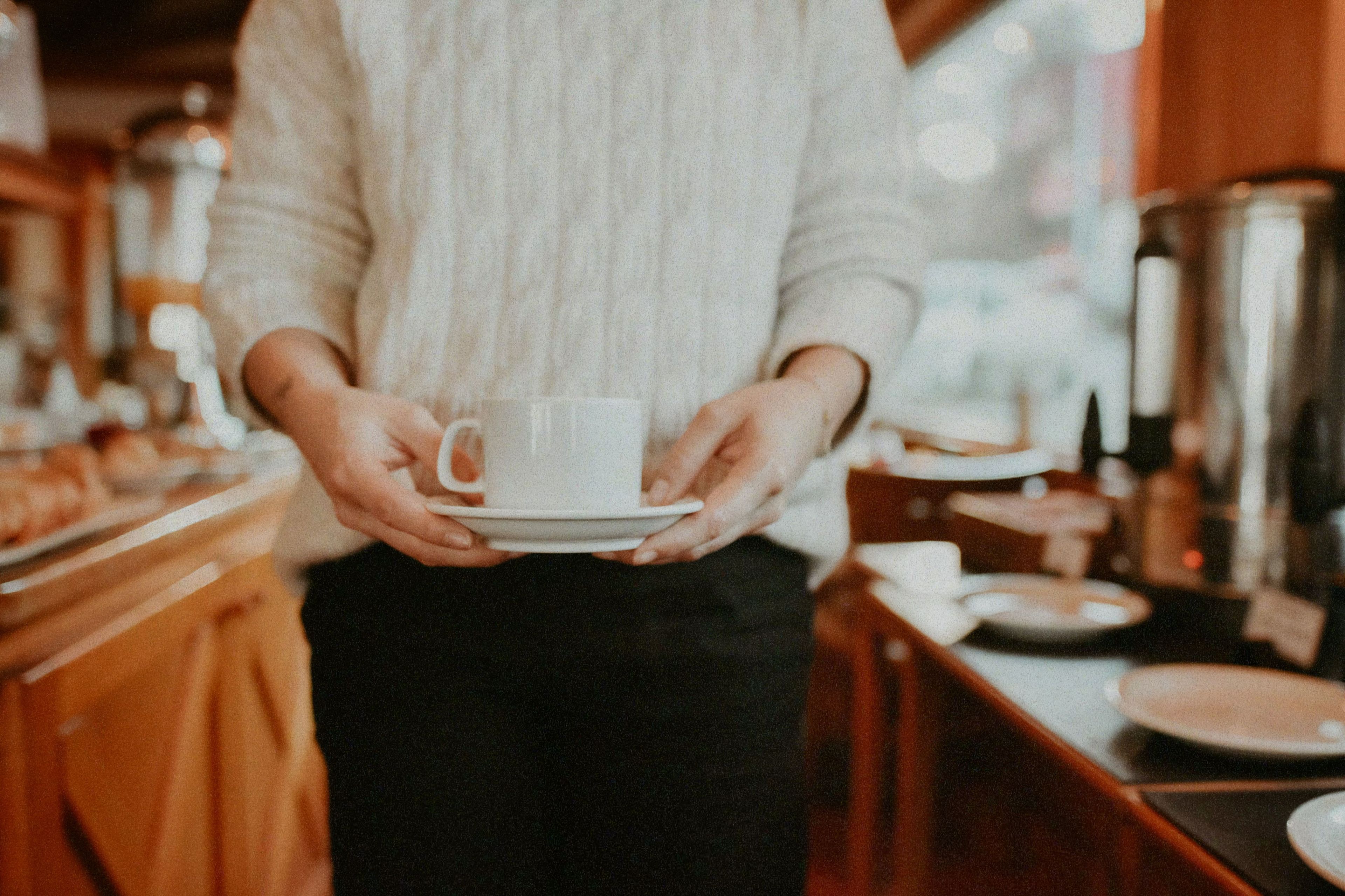 A person holding a white cup and saucer, likely in a cafe or restaurant setting.