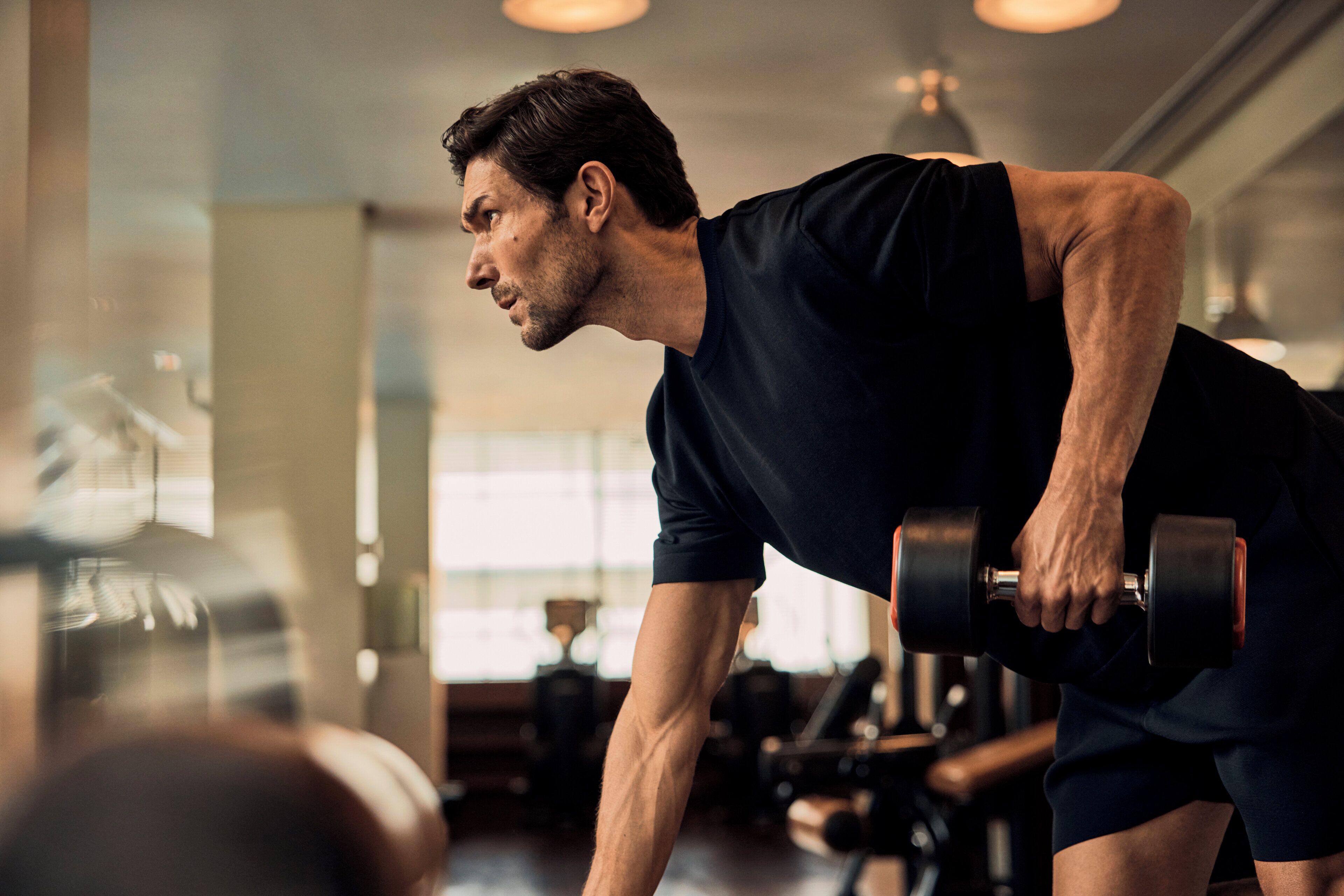 A person exercising with dumbbells in a gym with visible equipment and windows.