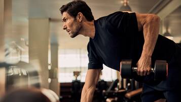 A person exercising with dumbbells in a gym with visible equipment and windows.