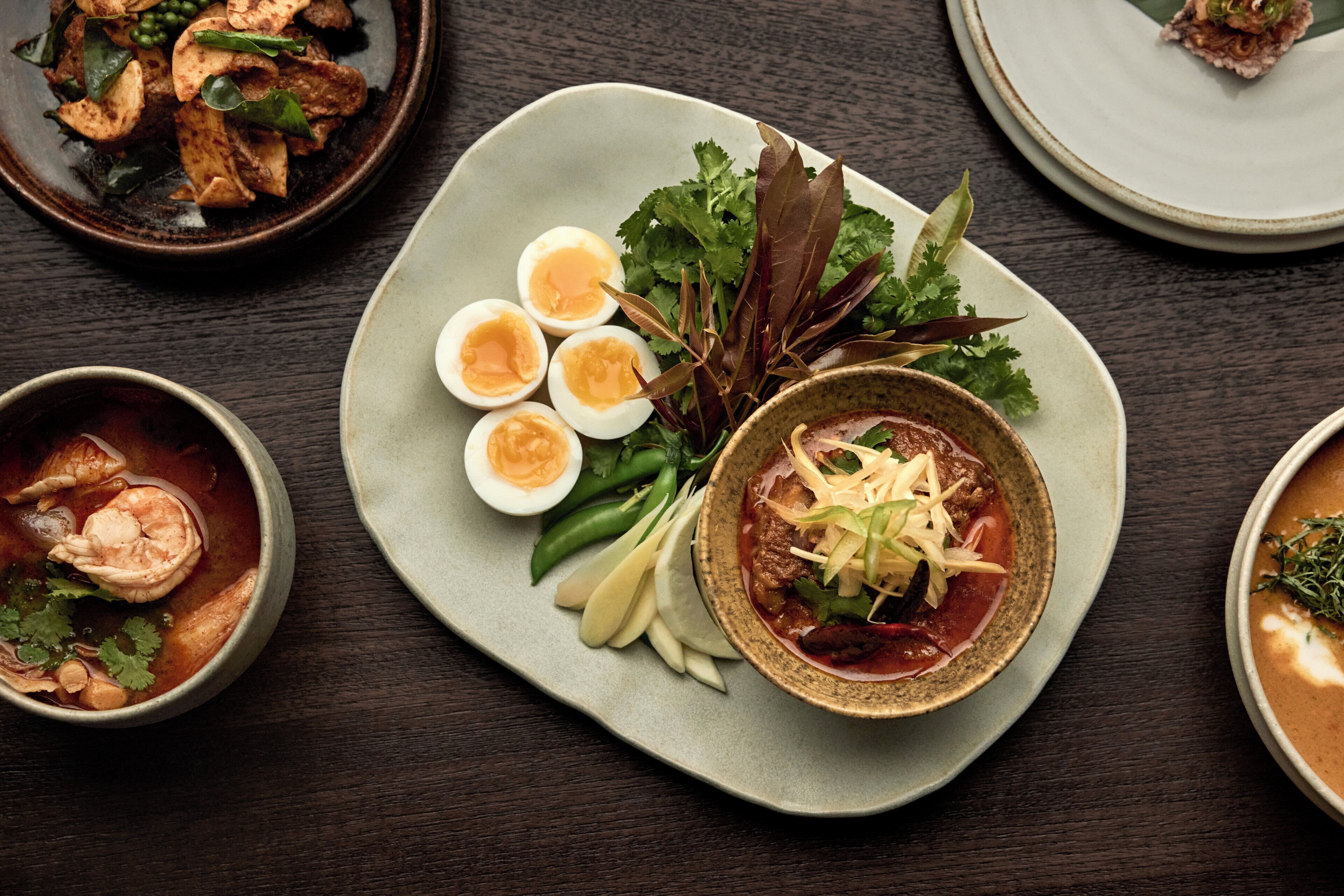Una mesa de comedor con diversos platos, entre ellos un tazón de sopa, un plato con carne y verduras en rodajas, y un bol de arroz.
