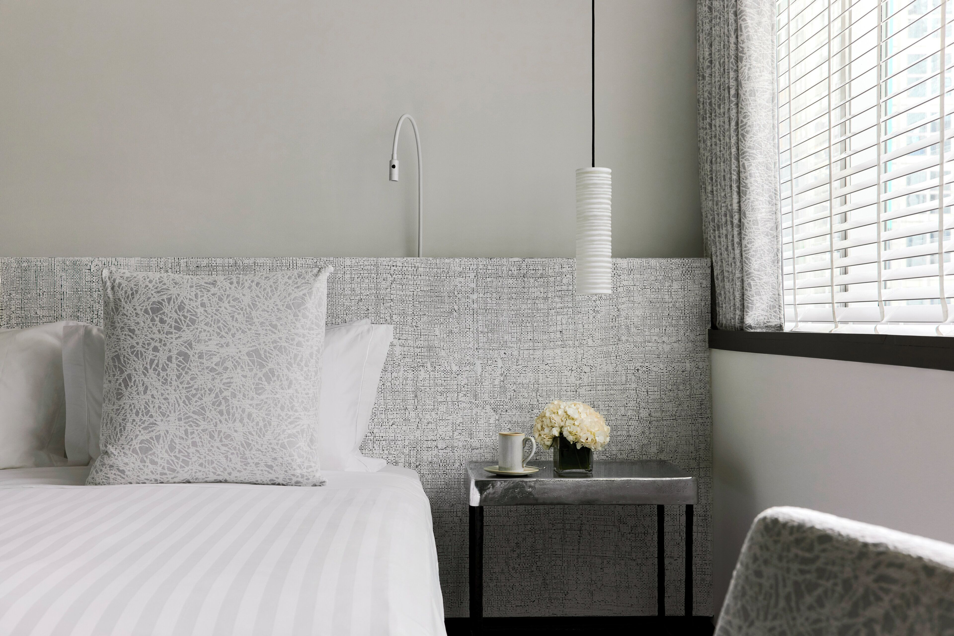 A hotel room with a bed, a nightstand with a vase of flowers, a hanging light, and a window with blinds.