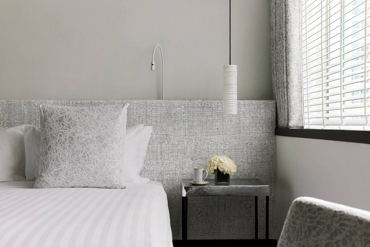 A hotel room with a bed, a nightstand with a vase of flowers, a hanging light, and a window with blinds.