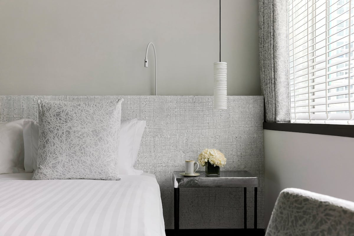 A hotel room with a bed, a nightstand with a vase of flowers, a hanging light, and a window with blinds.